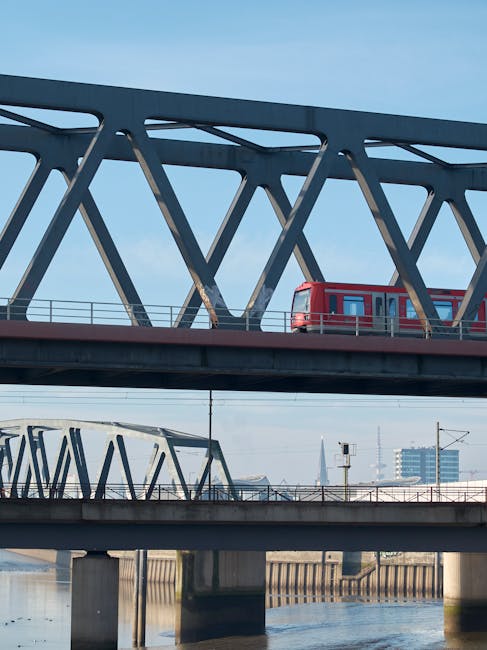 A red London Underground train travels across a modern, steel truss bridge with multiple intersecting beams, situated above a river. The scene is set in daylight with a clear blue sky, and the bridge�