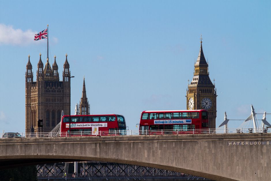 Two red double-decker buses traveling across Westminster Bridge in central London, with the iconic Big Ben clock tower and the Houses of Parliament visible in the background under a clear blue sky. Th